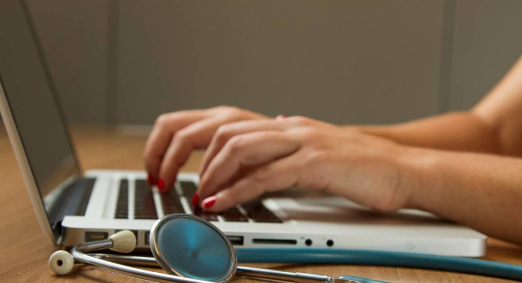 Mãos com unhas pintadas de vermelho digitando no teclado de um notebook, com um estetoscópio azul em primeiro plano sobre uma mesa de madeira.